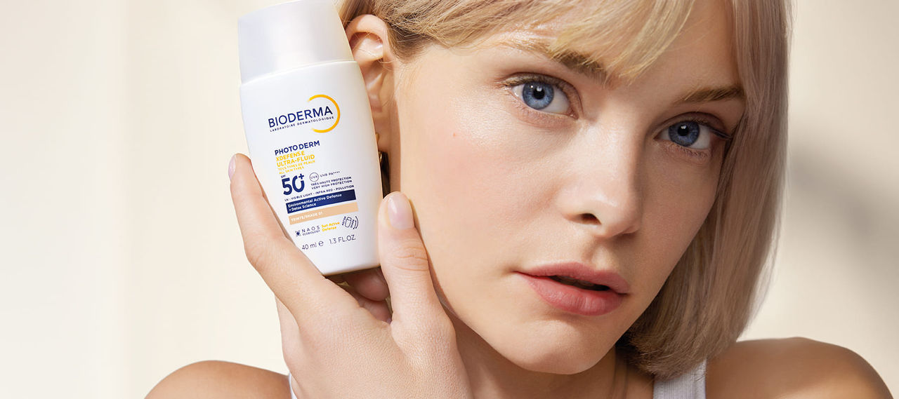 Mujer de piel clara con cabello rubio sostiene Photoderm XDefense SPF 50+ de BIODERMA cerca de su rostro, mostrando una piel protegida, luminosa y saludable.